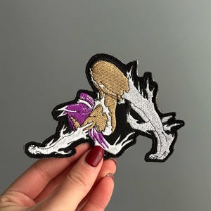 May include: An embroidered patch featuring a stylized figure with purple hair, tan skin, and white, jagged, flame-like accents. The design is outlined in black thread, creating a sharp contrast against the interior colors and the dark background.