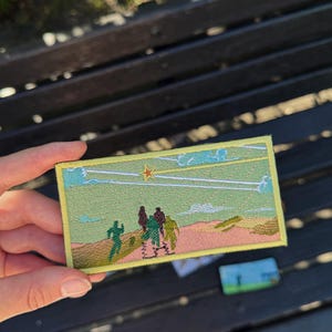 Puede incluir: Un parche rectangular bordado que presenta una escena de paisaje con un cielo verde claro, una estrella amarilla brillante y contornos de nubes blancas. El primer plano muestra un terreno desértico rosa con cuatro figuras oscuras y una corriendo.