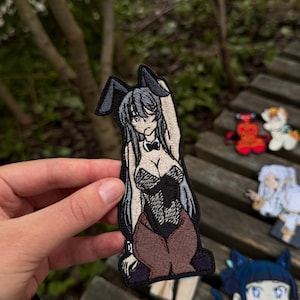 May include: An embroidered patch featuring an anime-style character with long grey hair and black bunny ears. The character wears a black leotard with a white collar and bow tie, paired with brown tights, depicted in a pose with one arm raised.