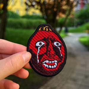 May include: A circular embroidered patch featuring a red, stylised face with a distressed expression, wide white eyes with black pupils, tears streaming down the cheeks, and a mouth showing white teeth, all outlined with a thick black border.