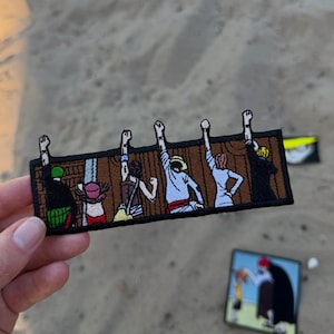 May include: A rectangular embroidered patch featuring six figures standing with their backs turned, each raising one arm with a fist in the air. The figures are positioned against a brown wooden wall background, outlined in a thick black border.