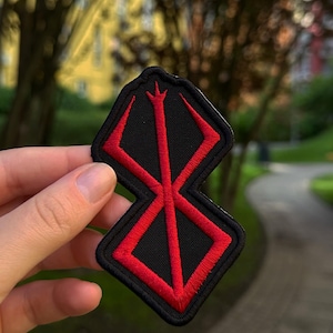Peut inclure: Un écusson brodé noir présentant un symbole géométrique rouge audacieux aux lignes nettes et angulaires avec une pointe verticale centrale. Le motif est entouré d'une bordure noire, créant un contraste marqué entre l'emblème rouge et le tissu noir.