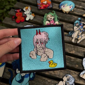 Op de afbeelding: Een vierkante geborduurde patch met een personage met roze haar, rode hoorns en rode ogen tegen een lichtblauwe achtergrond, met een kleine gele badeend in de rechterbenedenhoek, alles omrand door een dikke zwarte rand.