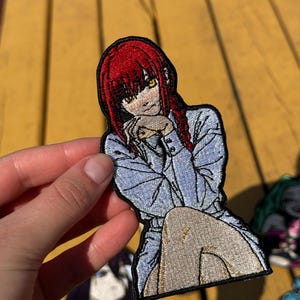 Può includere: Una toppa ricamata raffigurante un personaggio in stile anime con capelli rosso vivo, occhi gialli e una camicia azzurra. Il personaggio è raffigurato in posizione seduta con le mani vicino al mento, delineato da un bordo nero spesso.