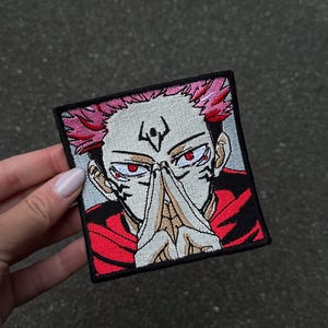 Könnte beinhalten: Ein quadratischer Stickaufnäher mit einem stilisierten Anime-Charakter mit stacheligen rosa Haaren, blasser Haut und intensiven roten Augen. Die Figur hat schwarze Gesichtsmarkierungen, trägt ein rotes Kleidungsstück und hält die Hände gefaltet.