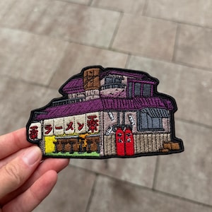 Puede incluir: Un parche bordado que representa un edificio con techo morado, una sección interior amarilla con taburetes de bar y dos puertas de color rojo brillante. La fachada presenta texto en japonés con caracteres rojos, una chimenea marrón y una linterna blanca.