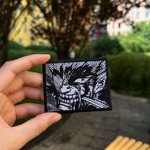 May include: A rectangular embroidered patch featuring a high-contrast black and white graphic illustration of a snarling character with sharp teeth and intense eyes. The design is bordered by a thick black stitched edge on a fabric base.