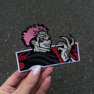 May include: An embroidered rectangular patch featuring a character with spiky pink hair, dark markings on the face, and sharp claws. The character is set against a red and black background, framed by a white border, showcasing detailed stitching.