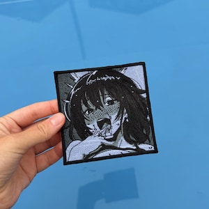 May include: A square embroidered patch featuring a black and white manga-style illustration of a character with dark hair, flushed cheeks, and an open mouth, framed by a thick black border with a light blue background.