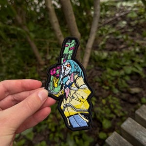 May include: An embroidered patch featuring a stylized character with light blue hair, wearing a yellow jacket over a white shirt and blue pants. The character holds a large green and pink firearm, with the entire design outlined in a thick black border.