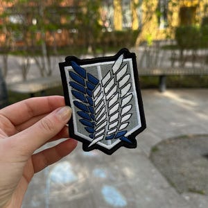 May include: A shield-shaped embroidered patch featuring the Wings of Freedom emblem. The design consists of a grey background with a thick black border, displaying two overlapping wings, one in dark blue and one in white, with intricate stitching.