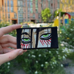 Pode incluir: Um emblema retangular bordado apresentando uma ilustração em close-up de dois olhos verdes intensos com mechas de cabelo escuro caindo sobre a testa e marcas verticais vermelhas abaixo dos olhos, tudo sobre um fundo preto sólido.