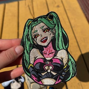 Op de afbeelding: Een geborduurde patch met een anime-stijl personage met lang groen haar, gele ogen en een brede grijns. Het personage draagt een roze en zwart topje met vetersluiting en heeft een roze nekgedeelte met rode markeringen, omlijnd met zwart garen.