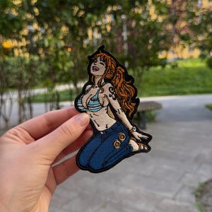 May include: An embroidered patch featuring a female anime character with long orange hair, wearing a light blue and white striped bikini top and dark blue jeans. The character is depicted in a kneeling pose with a black border outline.