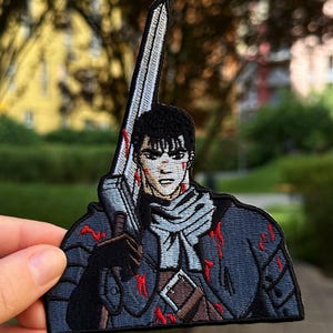 May include: An embroidered patch featuring a character with short black hair, a light gray scarf, and dark armor. The character holds a large silver sword upright. The design includes red accents representing blood splatters on the armor and face.