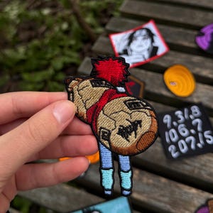 May include: An embroidered patch featuring a tan gourd with black line details and a red top section, positioned above two light blue legs. The design includes dark brown accents and a black border, showcasing a stylised character accessory.