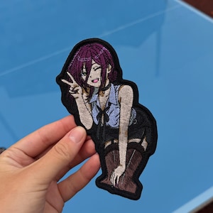 May include: An embroidered patch featuring an anime-style character with purple hair, wearing a light blue collared shirt, a black skirt, and brown boots. The character is making a peace sign gesture with one hand and has a black choker.