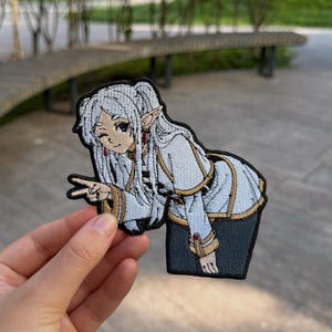Puede incluir: Un parche bordado que muestra un personaje de estilo anime con cabello largo blanco, orejas puntiagudas y una expresión de guiño. El personaje viste una chaqueta blanca y dorada con pantalones oscuros y hace un gesto de paz.