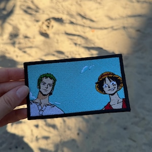 Op de afbeelding: Een rechthoekige geborduurde patch met een lichtblauwe achtergrond en twee anime-figuren. Links een figuur met kort groen haar in een wit overhemd. Rechts een figuur met een strooien hoed en een rood overhemd. De patch heeft een zwarte rand.