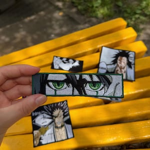 May include: A rectangular embroidered patch featuring a close-up of two intense green eyes with dark hair and a dark green border. Additional square patches with black and white anime-style character illustrations are visible in the background.