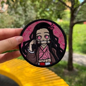 Può includere: Una toppa ricamata circolare raffigurante un personaggio con lunghi capelli neri, occhi rosa e un bavaglio di bambù. Il personaggio indossa un kimono rosa a motivi geometrici e un indumento marrone su sfondo rosa con piccoli fiori rosa.