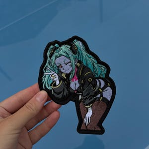 May include: An embroidered patch featuring a character with long teal hair, pale skin, and red eyes. The character wears a black jacket with yellow accents and dark clothing, making a peace sign with one hand while leaning forward.
