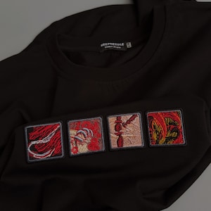 Puede incluir: Una camiseta negra con cuatro parches cuadrados bordados en el pecho. Cada parche muestra intrincados diseños abstractos en rojo, dorado y beige. La etiqueta interior del cuello dice KEEPNEEDLE con el texto Made in Ukraine.