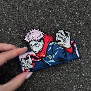 Pode incluir: Um emblema bordado retangular apresentando um personagem de anime estilizado com cabelo rosa espetado, vestindo uma jaqueta azul-escura com capuz vermelho. O personagem está em uma pose dinâmica com as mãos levantadas sobre um fundo rosa claro.
