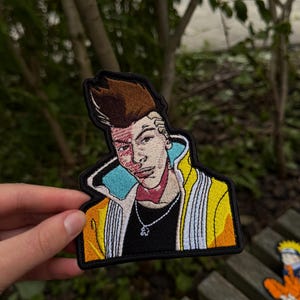 May include: An embroidered patch featuring a stylised character with brown hair, a red facial marking, and a yellow jacket with white accents. The character wears a black shirt and a silver chain necklace with a small pendant against a black border.