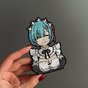 May include: An embroidered patch featuring an anime character with short light blue hair, a white maid outfit with ruffles, a black bow at the neck, and a white headpiece. The character has a smiling expression and is depicted from the chest up.