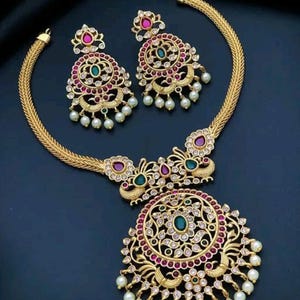 May include: A gold-coloured jewellery set comprising a necklace and matching earrings. The necklace features a large pendant with a central green stone, surrounded by red and clear stones. The earrings and pendant are adorned with faux pearls.