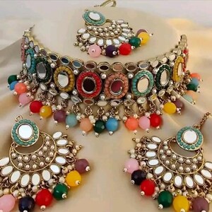 Puede incluir: Un conjunto de joyas dorado con un collar gargantilla, pendientes y una pieza para la cabeza. El conjunto incluye cuentas de colores, detalles de espejo y detalles perlados. El collar y los pendientes tienen forma de media luna.