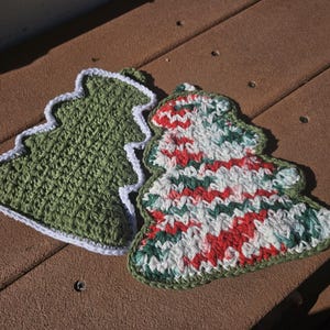 May include: Two handmade crochet Christmas tree-shaped potholders. One is solid green with a white border, the other features a red, green, and white pattern with a green border. Perfect for holiday decor.