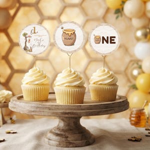 May include: Three cupcakes with white frosting on a wooden stand, each topped with a circular paper topper. The toppers feature illustrations of a honey pot, the text "One", and "Noah's 1st Birthday" with a tree and small animal characters.