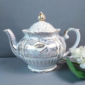 May include: White porcelain teapot with gold filigree and accents. The teapot features a curved spout, a loop handle, and a decorative lid with a gold finial. The design includes gold patterns and a diamond-shaped motif. A classic tea set item.