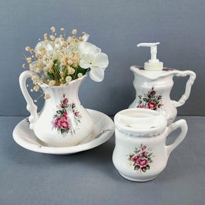 May include: A white ceramic bathroom set featuring a floral rose pattern. The set includes a pitcher on a matching basin, a soap dispenser with a white pump, and a toothbrush holder, all decorated with pink roses and green leaves on a white base.