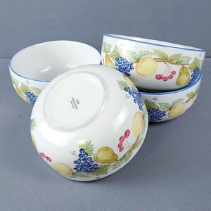 May include: Three white ceramic bowls featuring a painted fruit motif of blue grapes, yellow pears, and red cherries with green leaves. Each bowl has a thin blue rim. The bottom of one bowl is visible, showing the text RB BERNARDA PORTUGAL.
