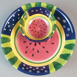 May include: A three-piece ceramic serving set featuring a watermelon design. The set includes a large plate, a medium plate, and a small bowl. The plates and bowl have a red watermelon slice design with black seeds, and are trimmed with blue, yellow, and green accents.