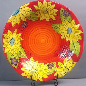 May include: A decorative ceramic plate with a vibrant sunflower design. The plate features a red rim and center with yellow sunflowers, green leaves, and purple accents. The plate is displayed on a black metal stand.