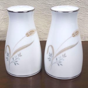 May include: A pair of white ceramic salt and pepper shakers with a wheat stalk design. The shakers have a silver rim at the top and a black rim at the base. The wheat stalks are tan and gray.