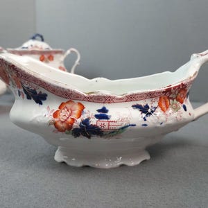 May include: White porcelain gravy boat with a floral and geometric pattern in red, orange, and blue. The boat has a handle and a scalloped rim. Other matching pieces are visible in the background.