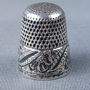 May include: A silver-colored metal thimble featuring a textured honeycomb pattern on the upper dome and a detailed embossed floral and leaf design around the base. The object has a metallic finish with darkened recessed areas for contrast.