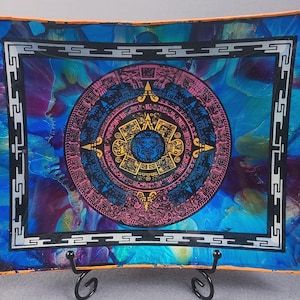 May include: Rectangular decorative glass tray with an Aztec calendar design. The tray has a vibrant blue, purple, and orange background. The calendar is in the center, with pink, yellow, and blue colors. The tray is supported by a black metal stand.