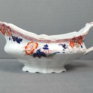 May include: White ceramic gravy boat with a decorative floral and geometric pattern in red, orange, and blue. The boat has a spout, a handle, and a scalloped base, showcasing an antique design.