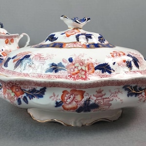 May include: A white porcelain serving dish with a lid, featuring a floral and pagoda design in shades of red, blue, and gold. The dish has handles and a scalloped edge, with a gold trim. A matching sugar bowl is visible in the background.