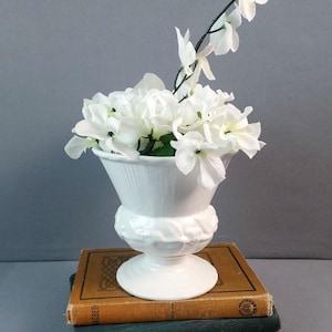 May include: A white ceramic vase filled with white flowers, resting on a stack of two antique books. The vase has a fluted top and a decorative base. The books have aged covers in shades of brown and green.