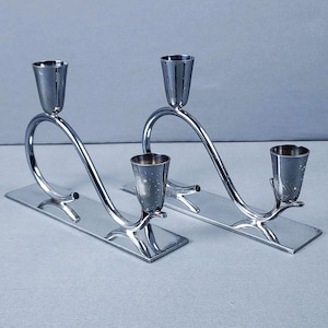 May include: A pair of silver-toned metal candle holders, each featuring a flat rectangular base and a curved, arching arm that supports two conical candle cups. The polished metallic surface reflects light, creating a sleek and uniform appearance.