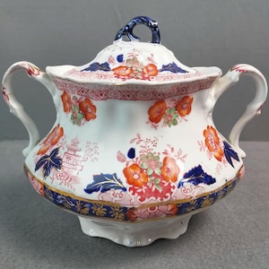 May include: A white ceramic sugar bowl with a lid, adorned with orange and pink floral patterns, blue accents, and gold trim. The bowl has two handles and a blue handle on the lid. The design includes a pagoda motif.