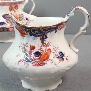 May include: White ceramic creamer with a decorative floral pattern in red, blue, and orange. The creamer has a curved handle and a scalloped rim with gold trim. A matching lid is visible in the background. The creamer is a vintage piece.