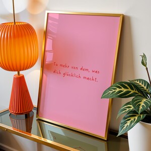 May include: A framed pink print with gold trim displays the text "Tu mehr von dem, was dich gl&uuml;cklich macht." An orange lamp with a ribbed shade and base sits on a glass-topped table. A potted plant with green and yellow leaves is also visible.